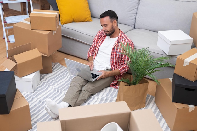 Un joven moreno se sienta con una computadora portátil entre cajas en un nuevo apartamento.