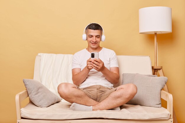 Un joven moreno sonriente con una camiseta blanca casual sentado en el sofá contra una pared beige navegando por Internet móvil en su teléfono inteligente y escuchando música en los auriculares