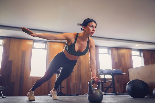 Joven motivada haciendo ejercicio de tabla con pesas rusas con una mano en el gimnasio