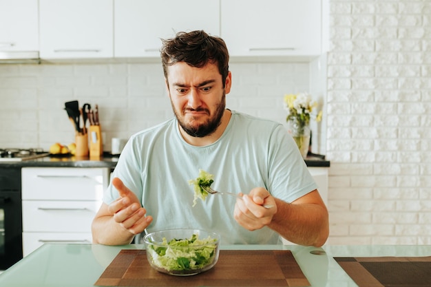 Un joven muestra la emoción de disgusto en su mano sosteniendo un tenedor con una jugosa ensalada verde un cuenco
