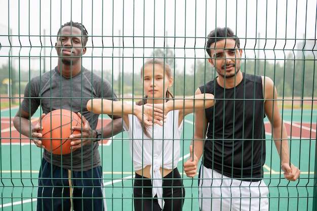 Joven mujer activa y dos hombres en ropa deportiva de pie junto a la red de baloncesto mientras descansan entre juegos