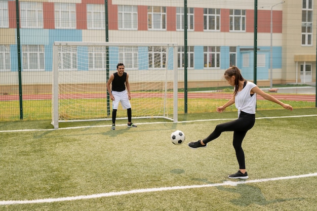 Joven mujer activa pateando un balón de fútbol hacia otro jugador de pie junto a la red en la puerta durante el juego en el campo