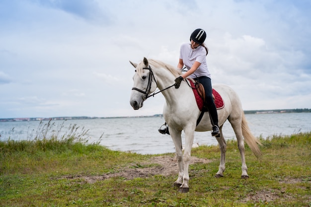 Joven mujer activa en traje ecuestre moviéndose a lo largo de la orilla del río mientras está sentado en la parte trasera del caballo de carreras blanco durante el entrenamiento