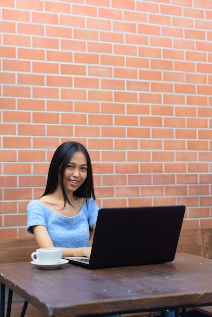 Joven mujer adolescente asiática feliz usando laptop con capuchino en mesa de madera