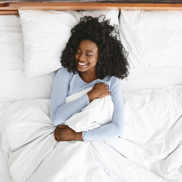 Joven mujer africana bien dormida despertando en la cama sonriendo y sosteniendo una manta con los ojos cerrados