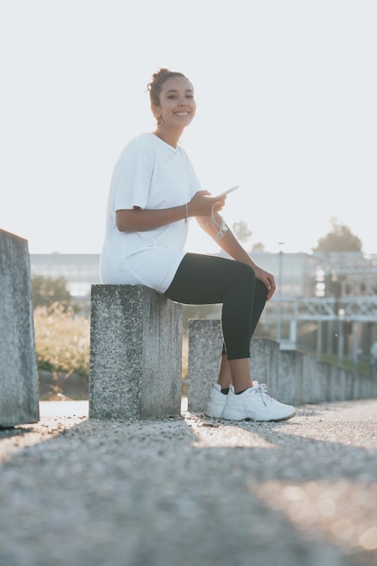 Joven mujer africana blanca gran sed comprobando el teléfono después de un día de ejercicio sentado cerca de la carretera