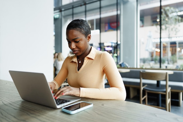 Joven mujer africana independiente que trabaja en una computadora portátil mientras está sentada en un coworking