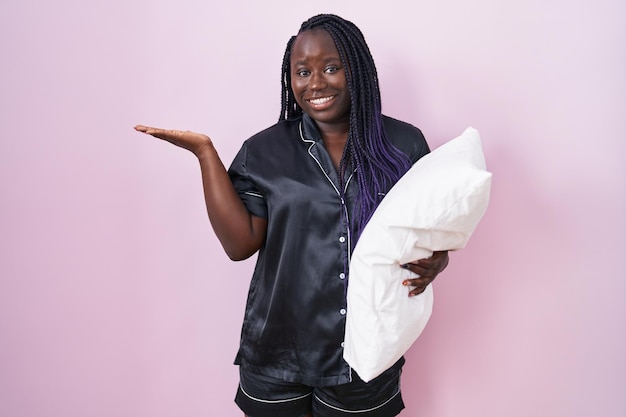 Joven mujer africana con pijama abrazando almohada sonriendo alegremente presentando y señalando con la palma de la mano mirando a la cámara