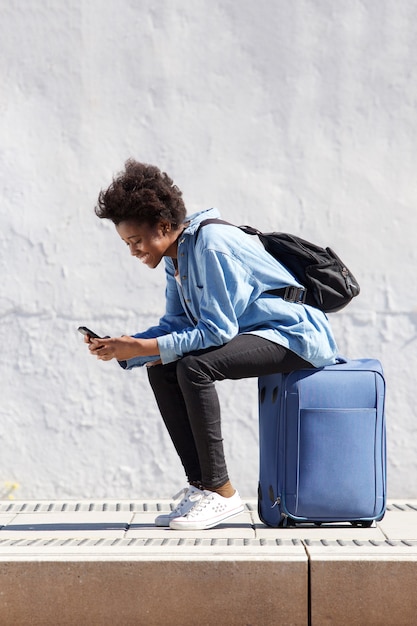 Joven mujer africana sentada en maleta con teléfono móvil