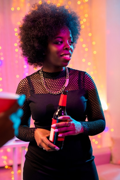 Joven mujer africana sonriente en traje negro sosteniendo una botella de cerveza mientras está de pie entre amigos y disfruta de la fiesta en el hogar.