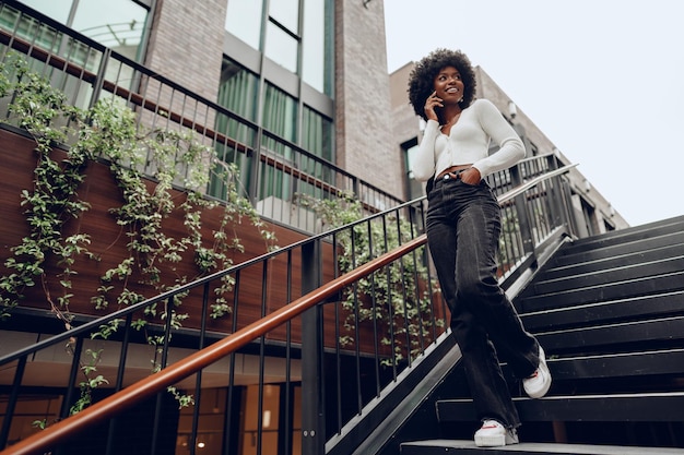 Joven mujer africana sonriente usando un teléfono inteligente bajando las escaleras en la ciudad