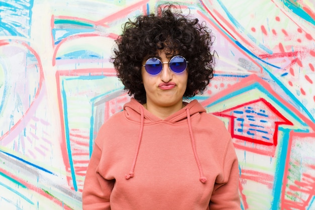 Joven mujer afro bonita que parece tonta y divertida con una expresión tonta con los ojos cruzados, bromeando y bromeando contra la pared de graffiti