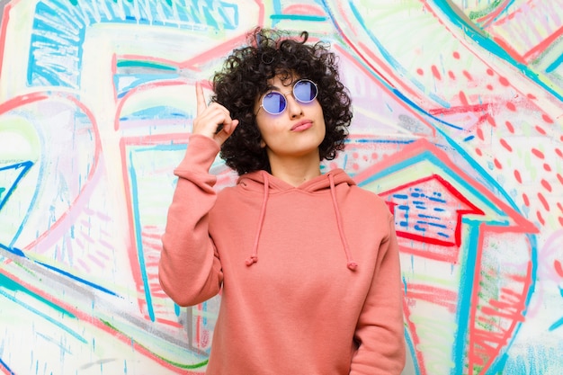 Joven mujer afro bonita que se siente como un genio sosteniendo el dedo con orgullo en el aire después de darse cuenta de una gran idea, diciendo eureka contra la pared de graffiti