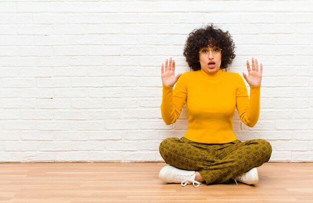 Joven mujer afro bonita que se siente estupefacta y asustada, temerosa de algo aterrador, con las manos abiertas al frente diciendo: aléjate sentado en el suelo