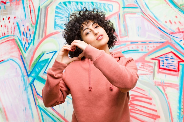 Joven mujer afro bonita sonriendo y sintiéndose feliz, linda, romántica y enamorada, haciendo forma de corazón con ambas manos contra la pared de graffiti