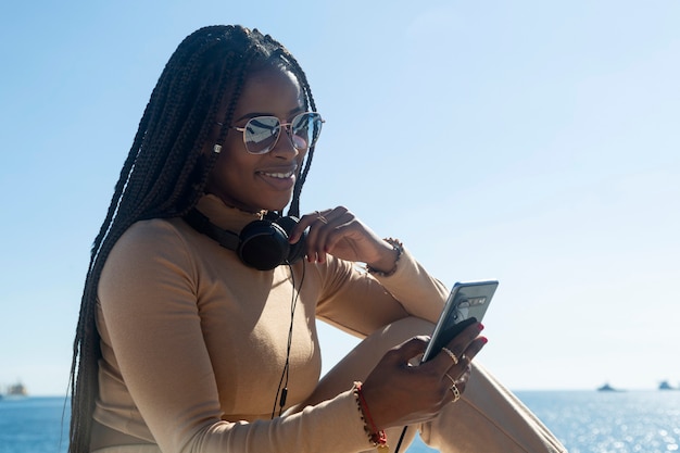 Joven mujer afro negra sonriendo mirando su teléfono celular, al aire libre con el cielo azul y el mar