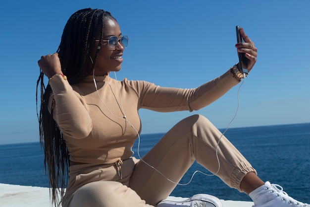 Joven mujer afro negra sonriendo mirando su teléfono celular, al aire libre con fondo de cielo azul y mar. Tecnología, comunicación, redes sociales.