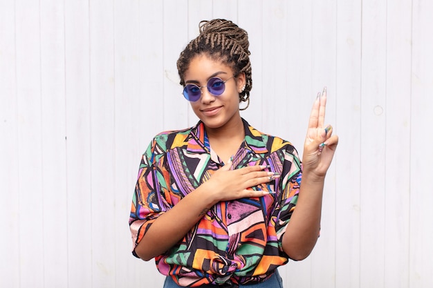 Joven mujer afro que parece feliz, segura y digna de confianza, sonriendo y mostrando el signo de la victoria, con una actitud positiva