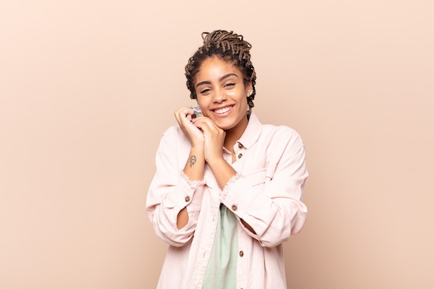 Joven mujer afro que se siente enamorada y se ve linda, adorable y feliz, sonriendo románticamente con las manos al lado de la cara