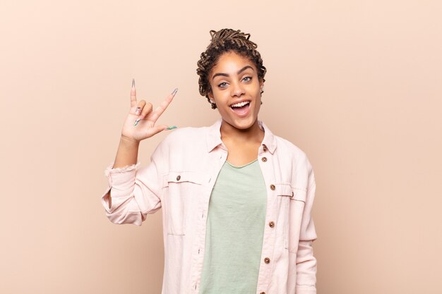 Joven mujer afro que se siente feliz, divertida, segura, positiva y rebelde, haciendo un letrero de rock o heavy metal con la mano