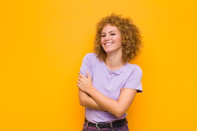 Joven mujer afro riendo alegremente con los brazos cruzados, con una pose relajada, positiva y satisfecha contra la pared naranja