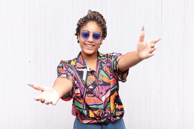 Joven mujer afro sonriendo alegremente dando un cálido, amistoso y amoroso abrazo de bienvenida, sintiéndose feliz y adorable