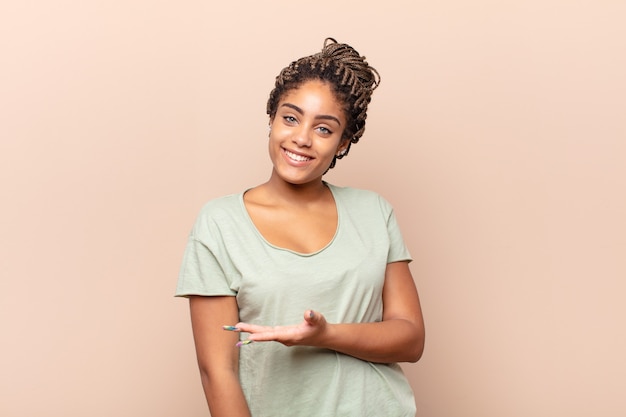 Joven mujer afro sonriendo alegremente, sintiéndose feliz y mostrando un concepto en el espacio de la copia con la palma de la mano