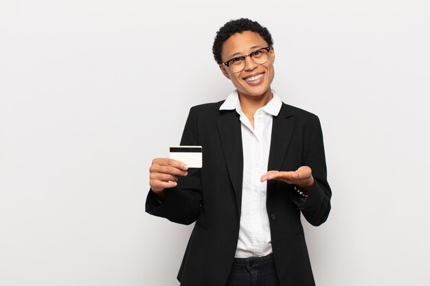 Joven mujer afro sonriendo alegremente, sintiéndose feliz y mostrando un concepto en el espacio de la copia con la palma de la mano