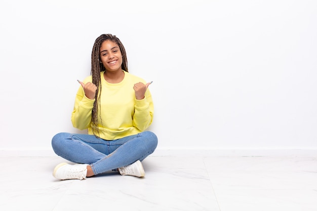 Joven mujer afro sonriendo con alegría y luciendo feliz, sintiéndose despreocupada y positiva con ambos pulgares hacia arriba