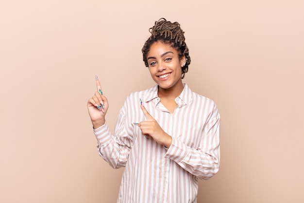 Foto joven mujer afro sonriendo felizmente y apuntando hacia un lado y hacia arriba con ambas manos mostrando el objeto en el espacio de la copia