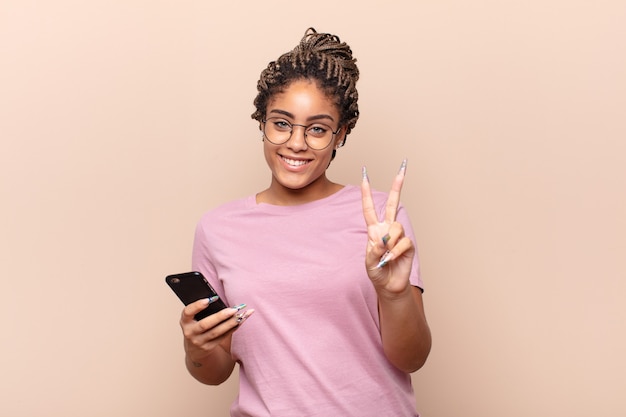 Joven mujer afro sonriendo y mirando amigable mostrando el número dos o el segundo con la mano hacia adelante contando el concepto de teléfono inteligente