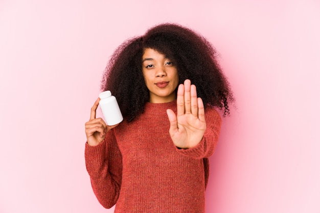 Joven mujer afro sosteniendo una botella de píldoras de vitaminas y de pie con la mano extendida que muestra la señal de stop