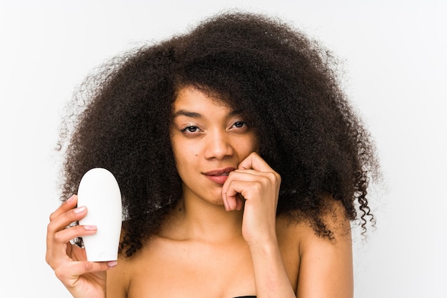 Joven mujer afro sosteniendo una crema hidratante aislada mordiéndose las uñas, nerviosa y muy ansiosa.