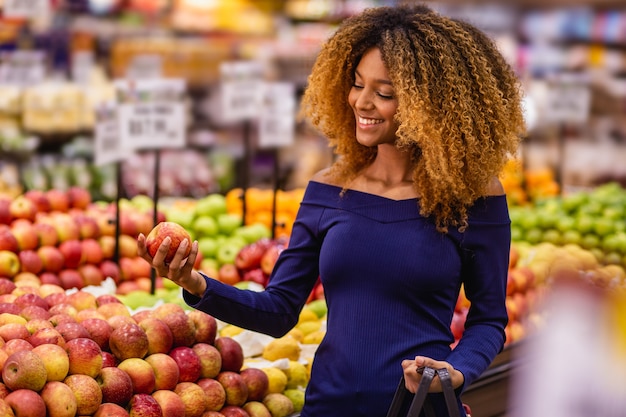 Joven mujer afro en supermercado comprando manzanas.