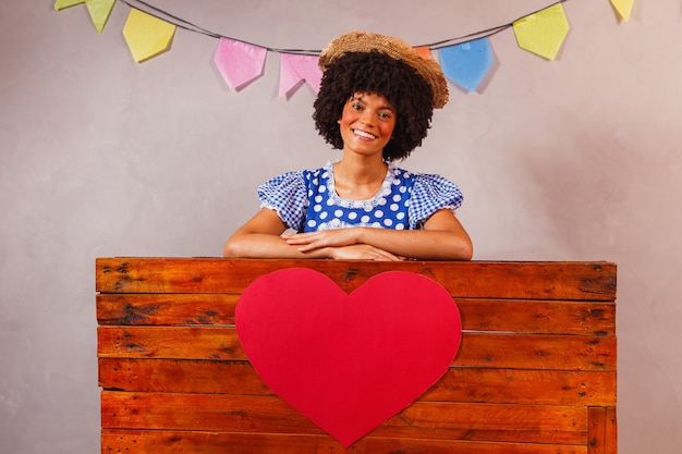 Joven mujer afro vestida para la fiesta de junina detrás de una tabla de madera con un corazón