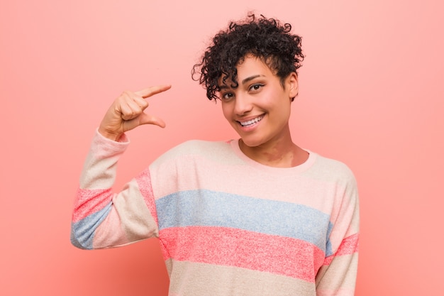 Joven mujer afroamericana adolescente mezclada sosteniendo algo poco con los dedos índice, sonriente y confiado.