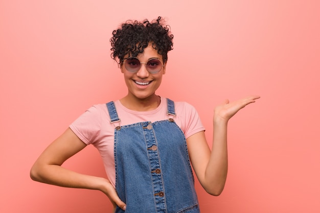 Joven mujer afroamericana adolescente mixta mostrando un espacio de copia en una palma y sosteniendo otra mano en la cintura.