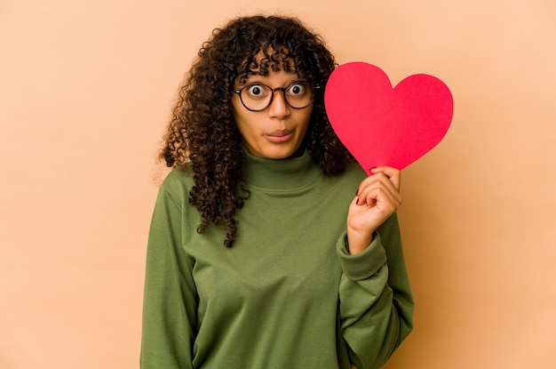 Joven mujer afroamericana afro sosteniendo un corazón de San Valentín se encoge de hombros y abre los ojos confundidos.