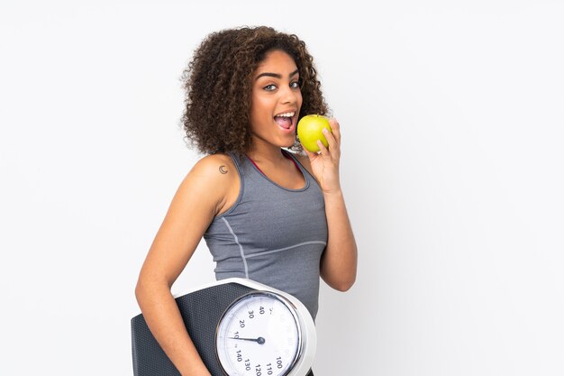 Joven mujer afroamericana aislada en blanco con máquina de pesaje y con una manzana