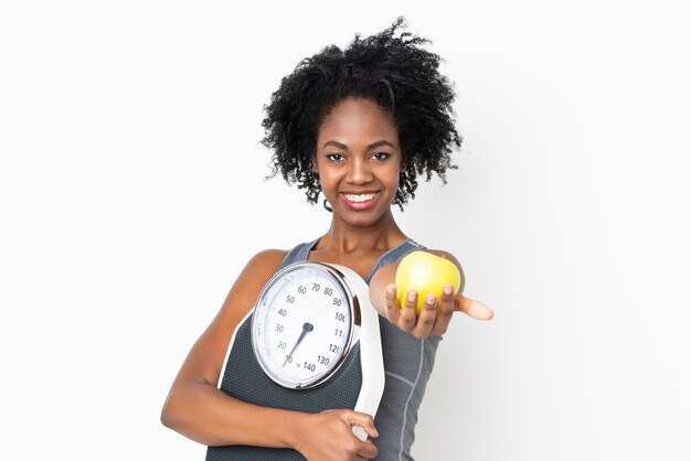 Joven mujer afroamericana aislada en la pared blanca con máquina de pesaje y ofreciendo una manzana