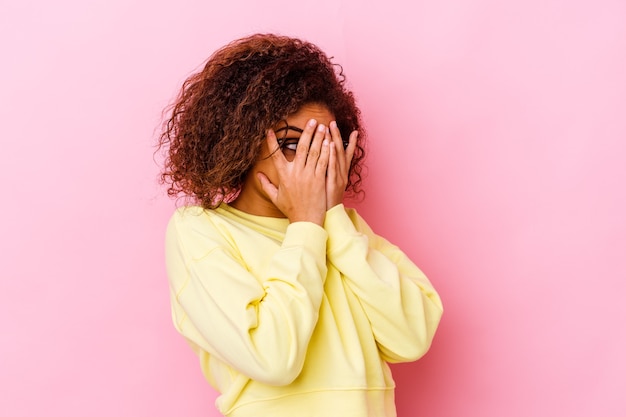 Joven mujer afroamericana aislada en la pared rosa parpadea a través de los dedos asustada y nerviosa.