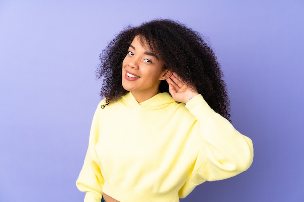 Joven mujer afroamericana aislada en púrpura escuchando algo poniendo la mano en la oreja