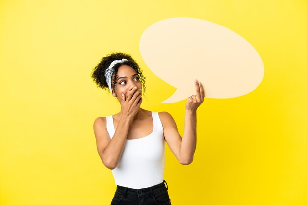 Joven mujer afroamericana aislada sobre fondo amarillo sosteniendo un bocadillo vacío con expresión de sorpresa