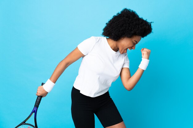 Joven mujer afroamericana aislada sobre fondo azul jugando tenis y celebrando una victoria