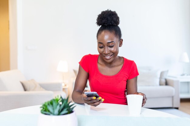 Joven mujer afroamericana bebiendo café y charlando con amigos en la red social con su teléfono móvil