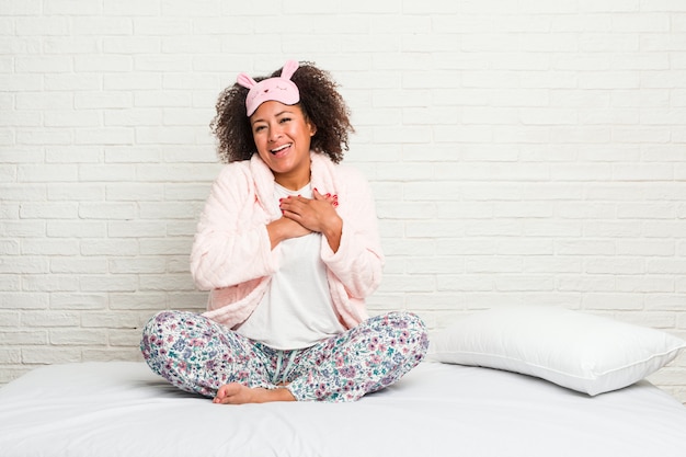 Joven mujer afroamericana en la cama con pijama tiene expresión amigable, presionando la palma contra el pecho. Concepto de amor