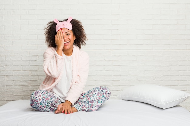 Joven mujer afroamericana en la cama vistiendo pijama divirtiéndose cubriendo la mitad de la cara con la palma.