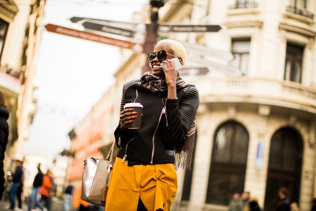 Joven mujer afroamericana caminando por la ciudad con café en mano y con teléfono móvil