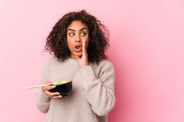 Joven mujer afroamericana comiendo fideos está diciendo una noticia secreta de frenado en caliente y mirando a un lado