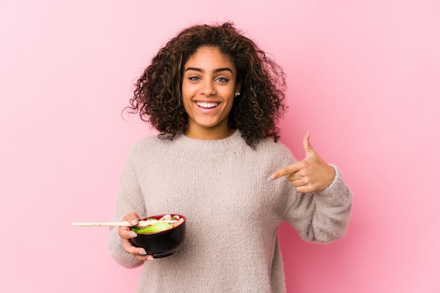 Joven mujer afroamericana comiendo fideos persona apuntando con la mano a una camisa espacio en blanco, orgulloso y confiado